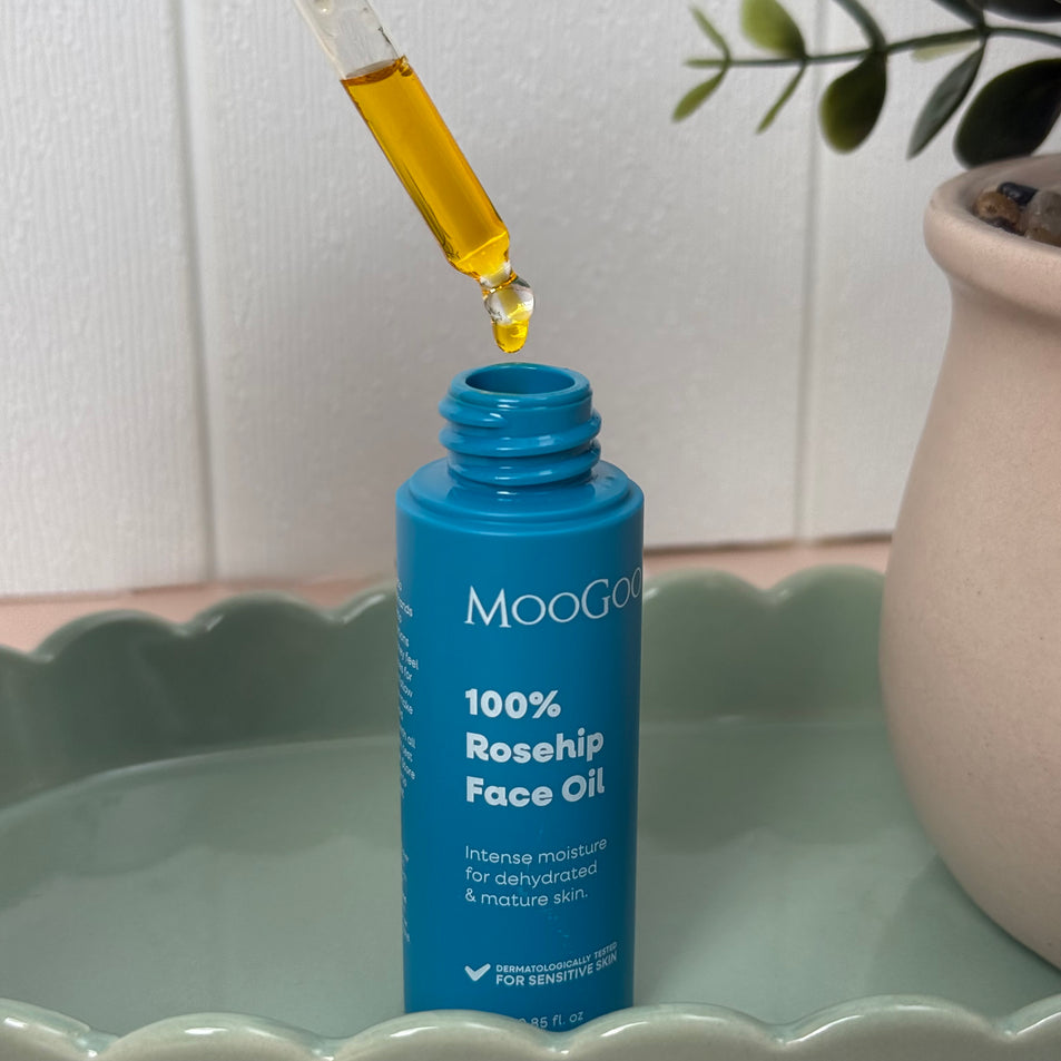 MooGoo 100% Rosehip Face Oil in a blue bottle on a sage green scalloped tray, with a glass dropper dispensing golden oil above the bottle. A potted plant sits in the background, creating a calming skincare setting.