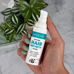 A hand holding a white pump bottle of MooGoo Natural Hair Growth Serum, with a green potted plant in the background on a light marble surface.