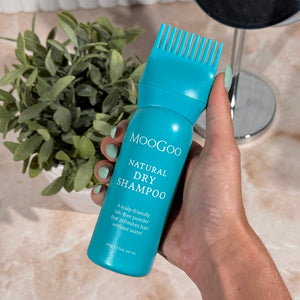 Hand holding a blue bottle of MooGoo Natural Dry Shampoo with a plant and tiles in the background.