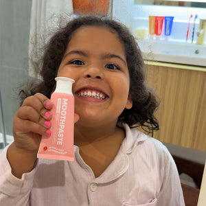 A young child smiling while holding a pink tube of MooGoo Strawberry Kids Moothpaste in their hand, with a bathroom background visible.