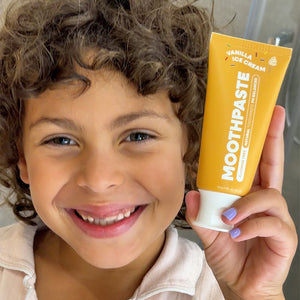A smiling child with curly brown hair holds up a tube of Vanilla Kids Moothpaste toothpaste. The tube is bright yellow with white text and a white cap, while the child is wearing a light beige top and has light purple painted fingernails.